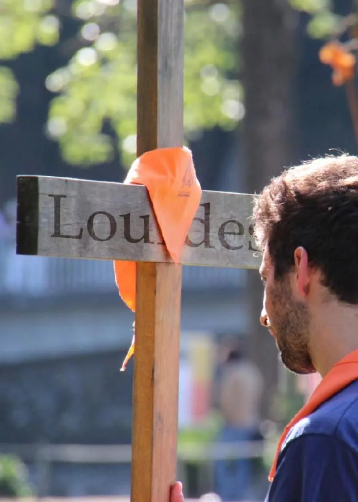 Un jove amb el mocador taronja davant d’una creu de fusta amb la paraula Lourdes